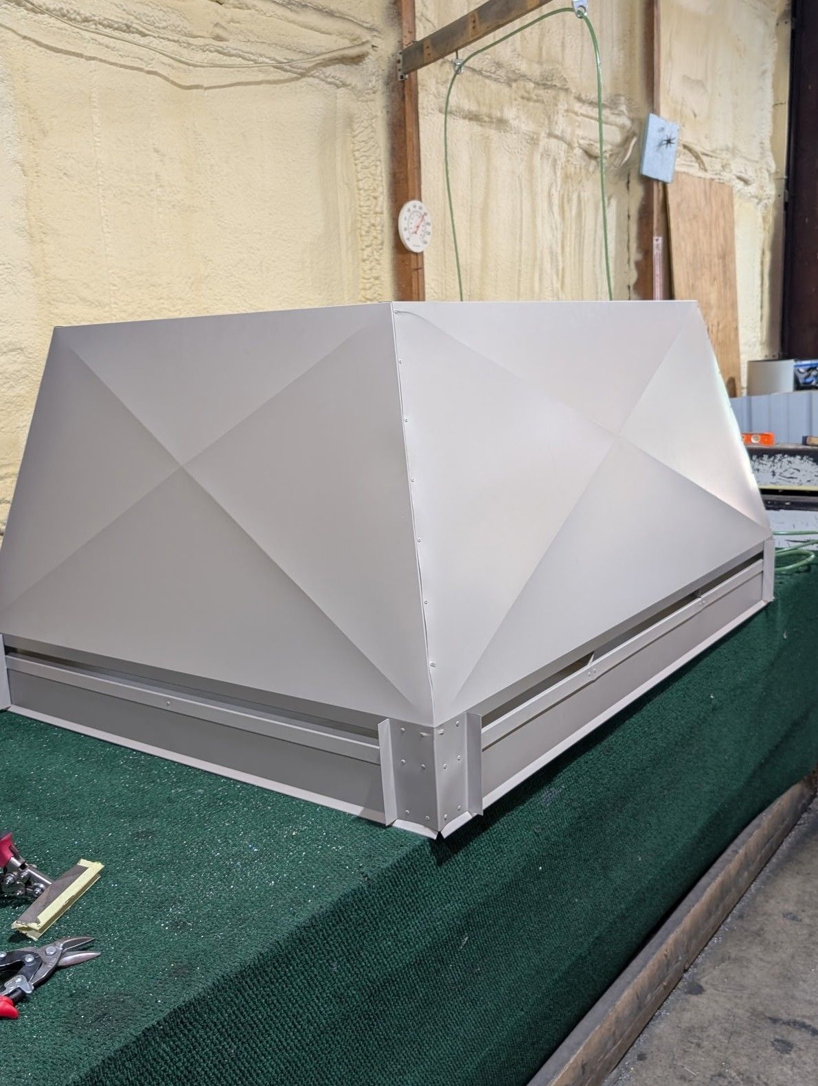 Gray faceted rooftop vent or HVAC unit on a green work surface, indoors near insulation paneling