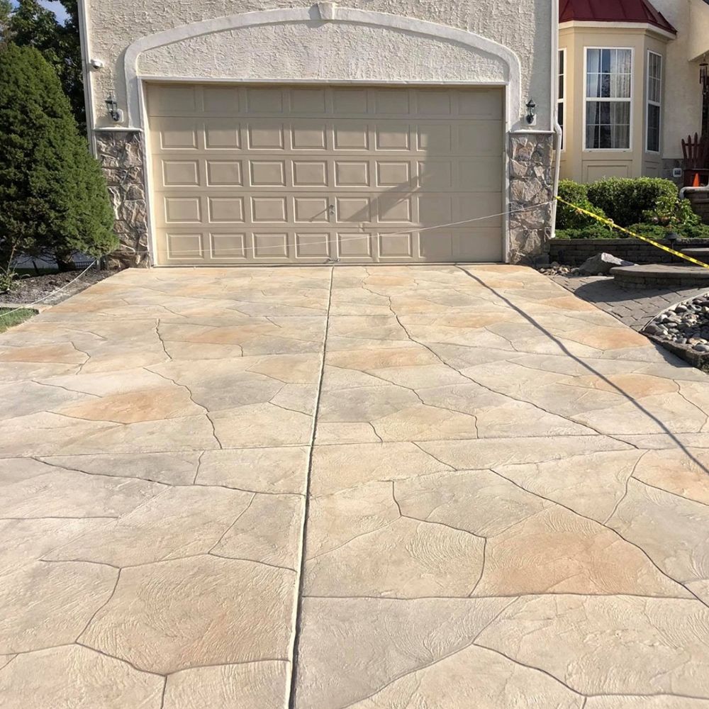 Driveway leading to a beige garage with stone accents and a large concrete patio in front.