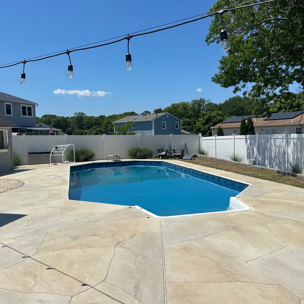 Backyard pool with blue water, concrete patio, string lights, and white fence under a clear sky