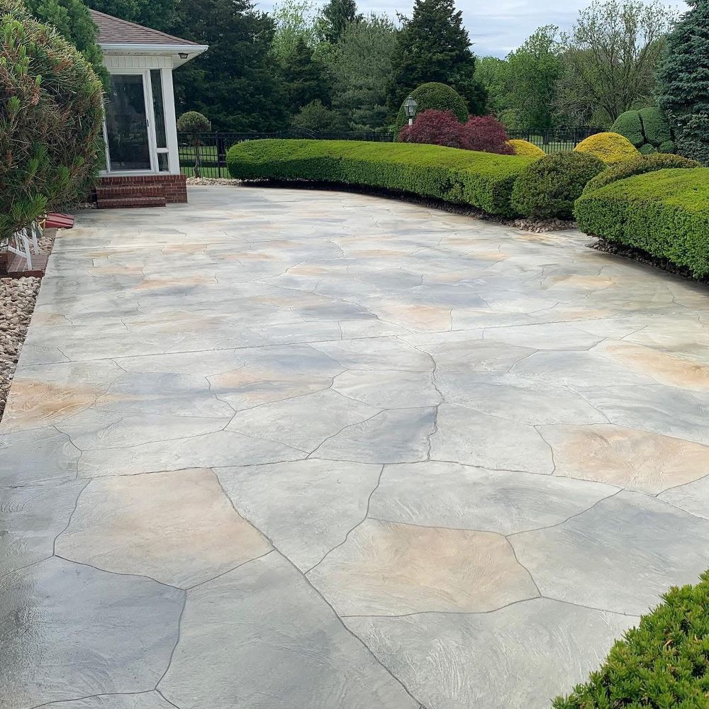 Curved stone patio beside a house, bordered by manicured shrubs and garden beds.