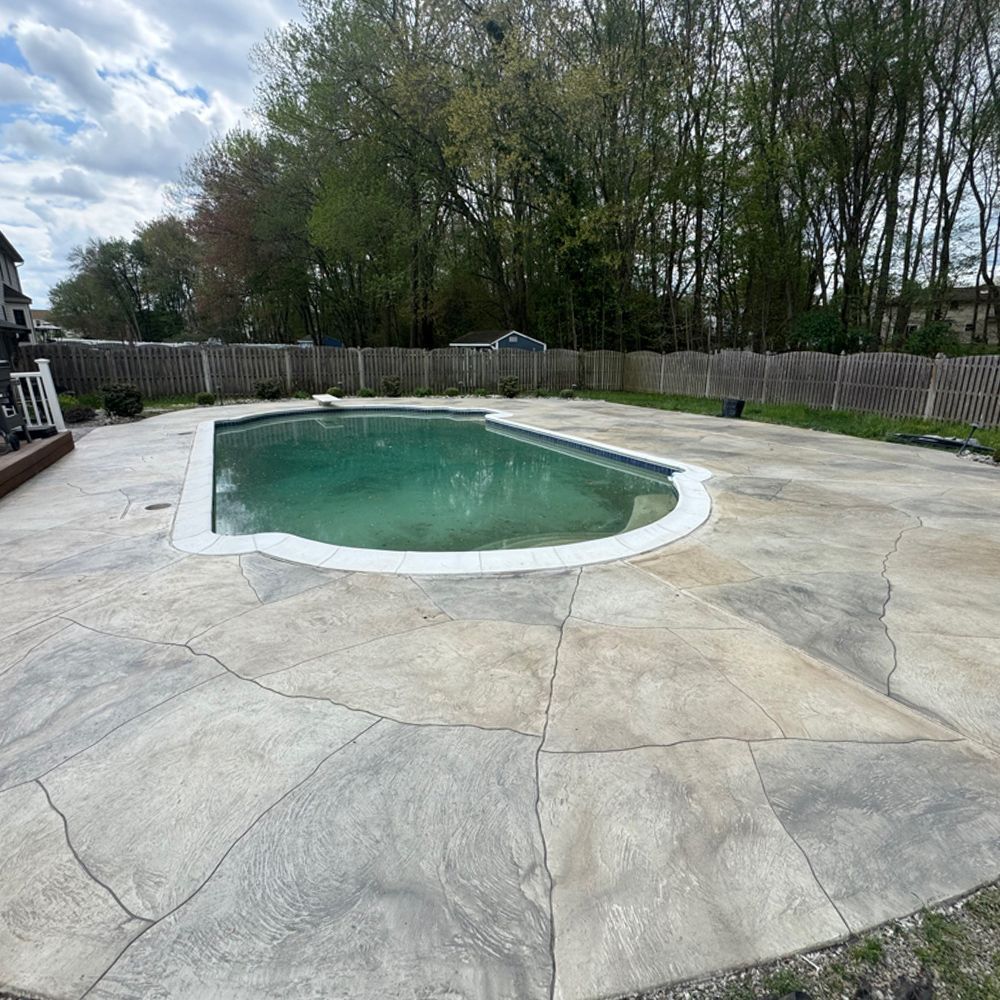 Empty backyard swimming pool with green water, surrounded by a cracked concrete patio and trees.