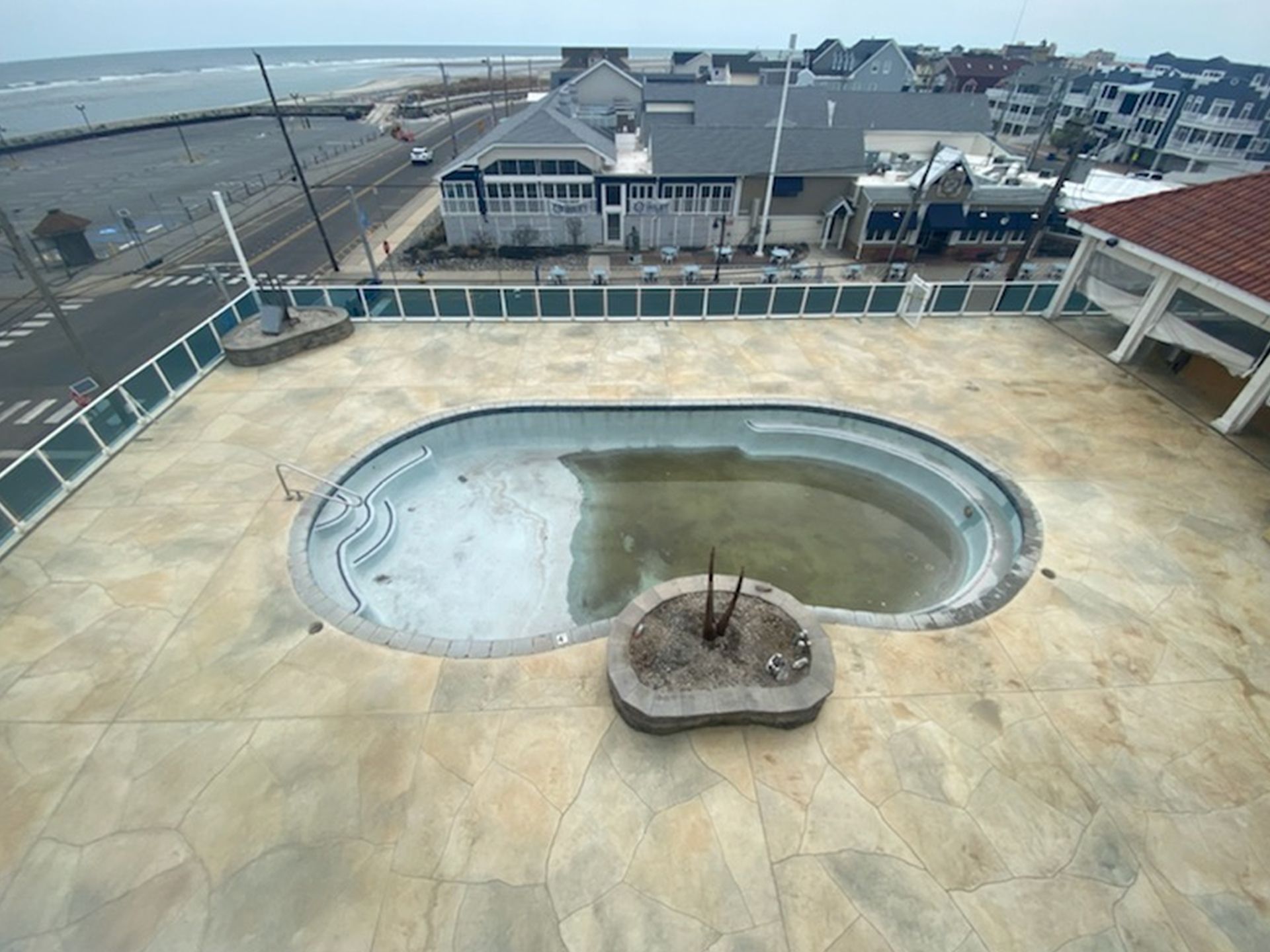 Empty drained rooftop pool beside a seaside boardwalk and buildings under a cloudy sky