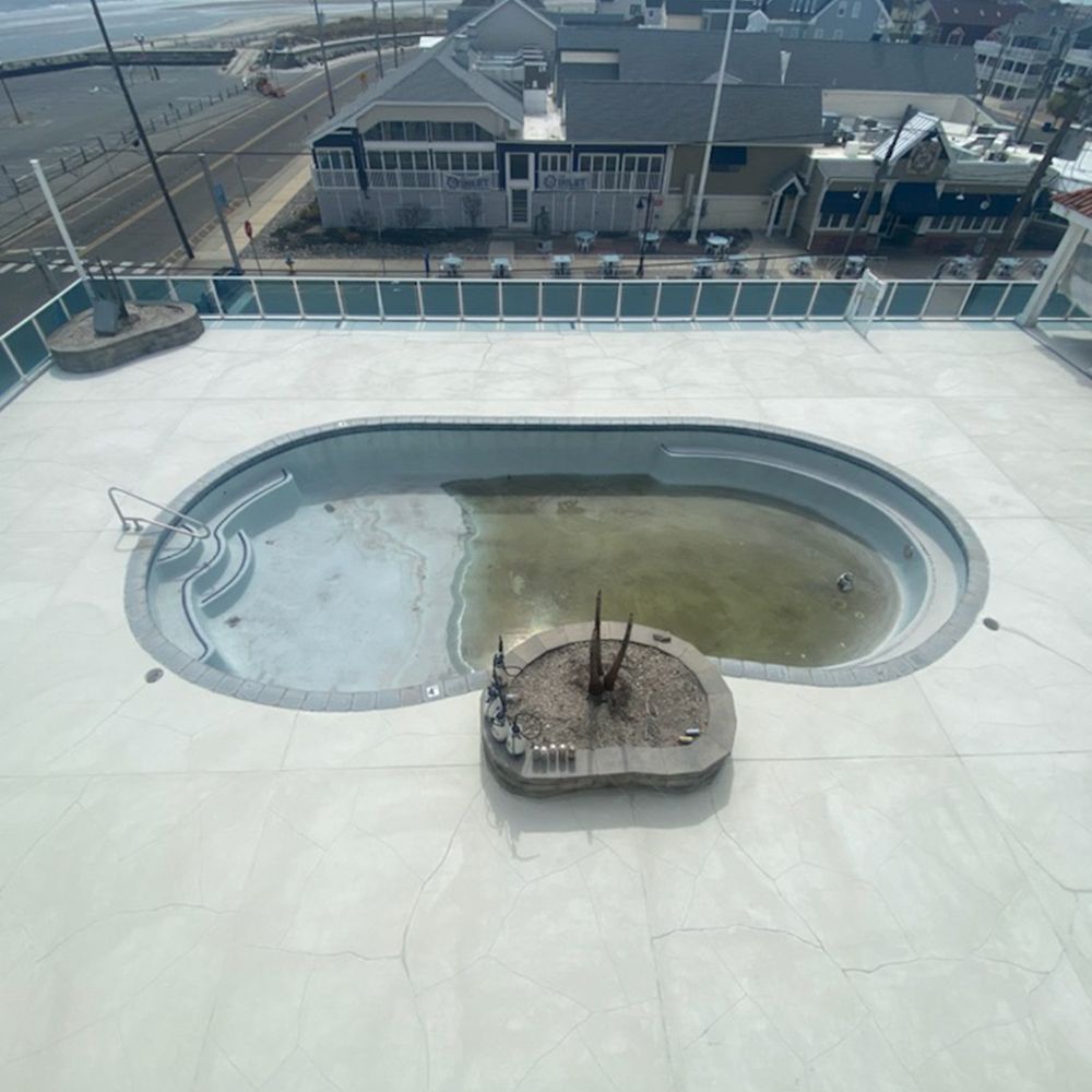 Empty rooftop pool with a shallow, dirty basin and a small central fire pit, overlooking industrial buildings.