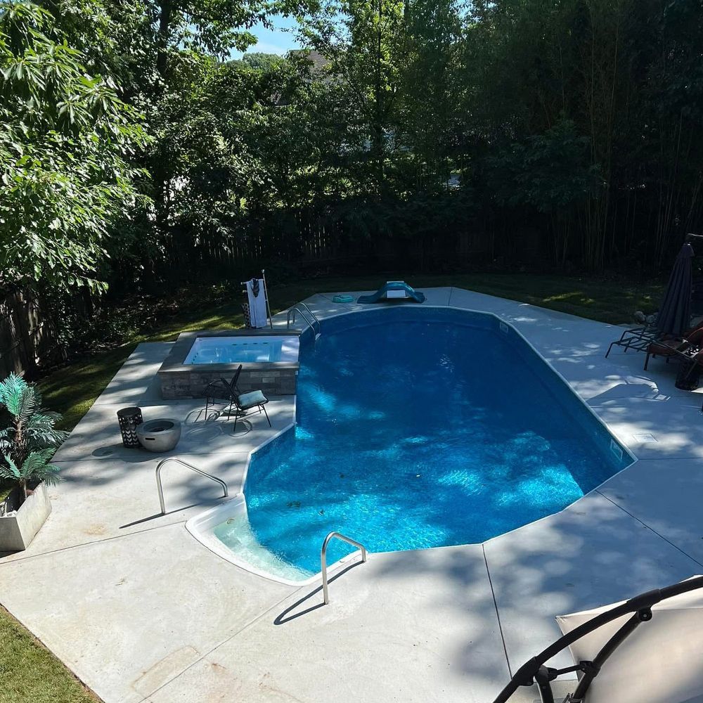 Backyard swimming pool with blue water, patio chairs, and surrounding trees in sunlight