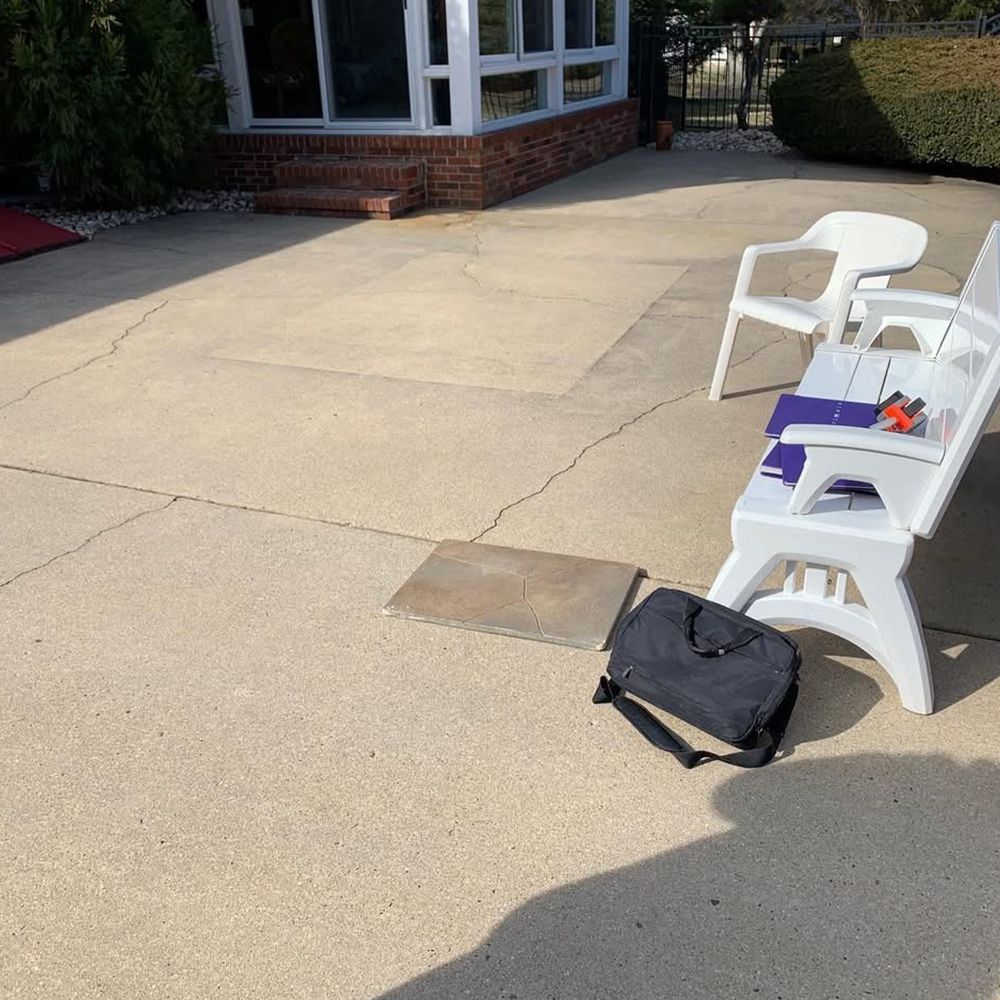 White folding chairs and a black bag on a sunlit concrete patio outside a brick building.