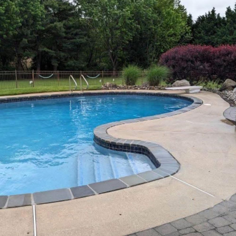 Curved backyard swimming pool with blue water, beige deck, and landscaped greenery