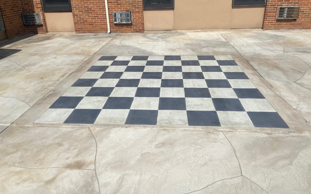 Outdoor concrete patio with a painted black-and-white checkerboard game board near a brick wall.