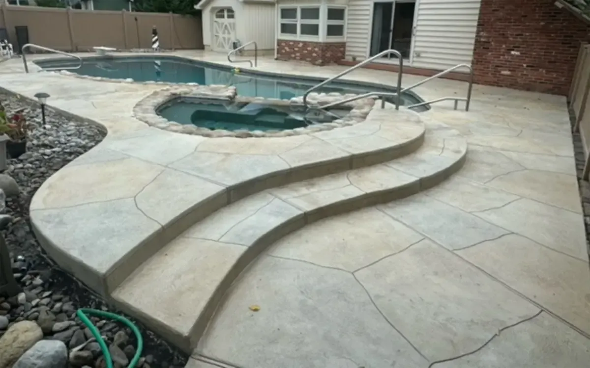 Curved concrete pool deck with built-in steps and a hot tub beside a house and brick wall