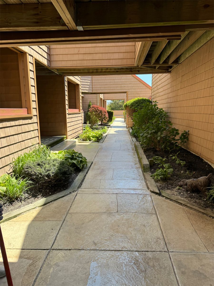 Covered apartment walkway with tan brick walls, concrete path, and small shrubs leading to sunlight