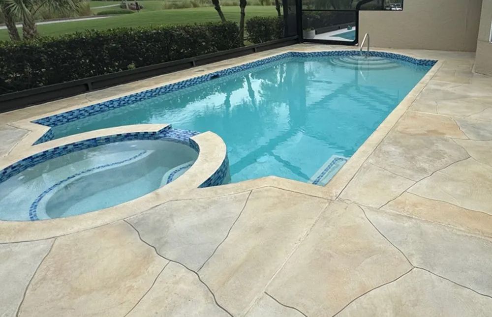 Backyard swimming pool with attached hot tub and beige stone patio beside green lawn.