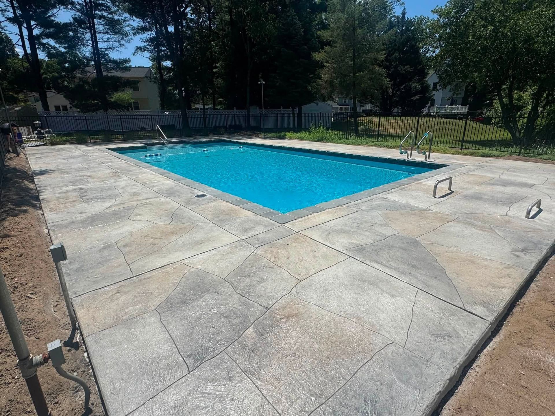 Rectangular outdoor swimming pool with blue water on a concrete deck, surrounded by trees and fencing.
