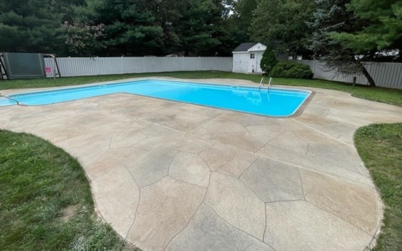 Backyard swimming pool with a tan stamped concrete deck and surrounding fence, trees, and shed