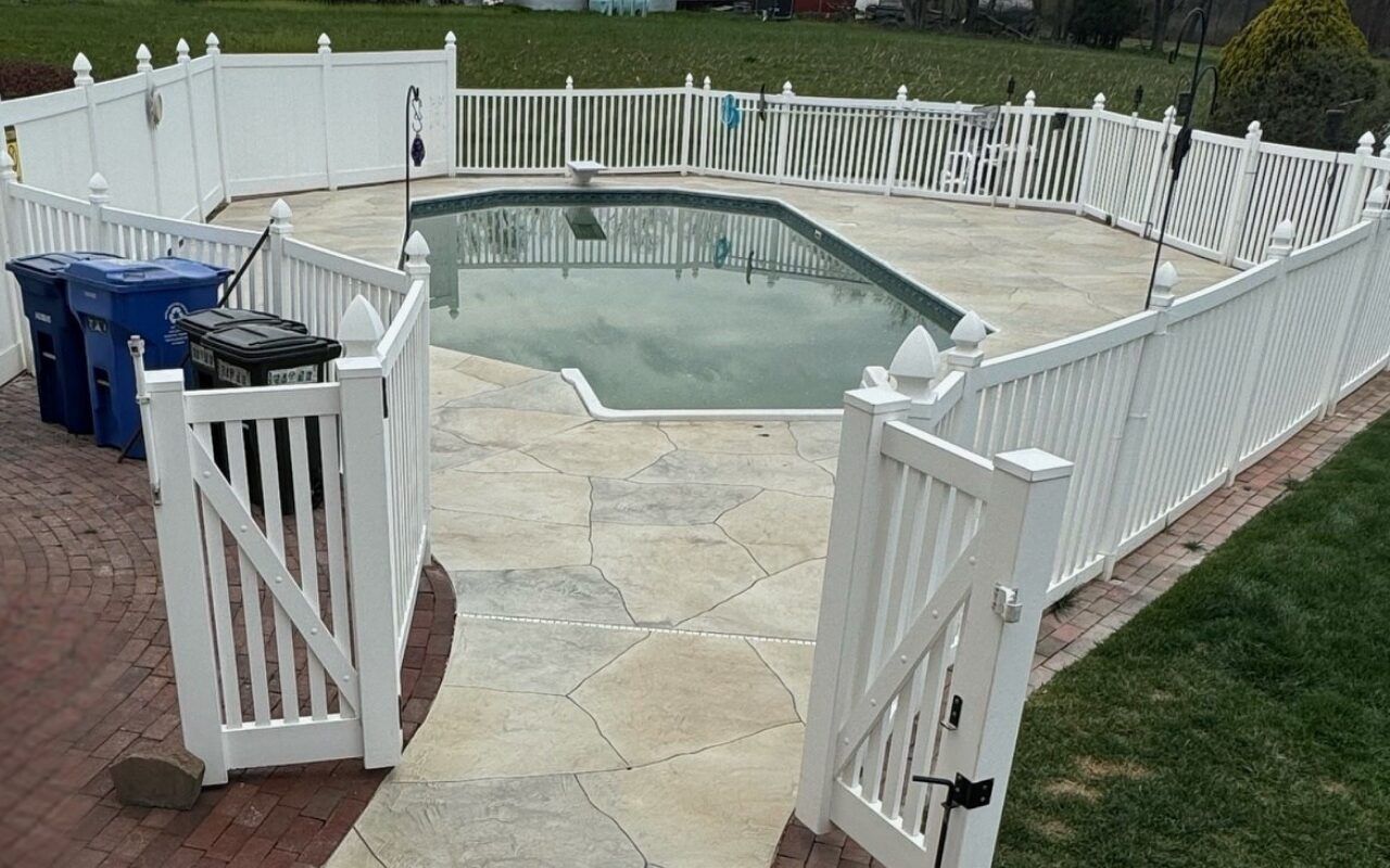 Fenced-in backyard pool with white gate, stone patio, and blue trash bins nearby
