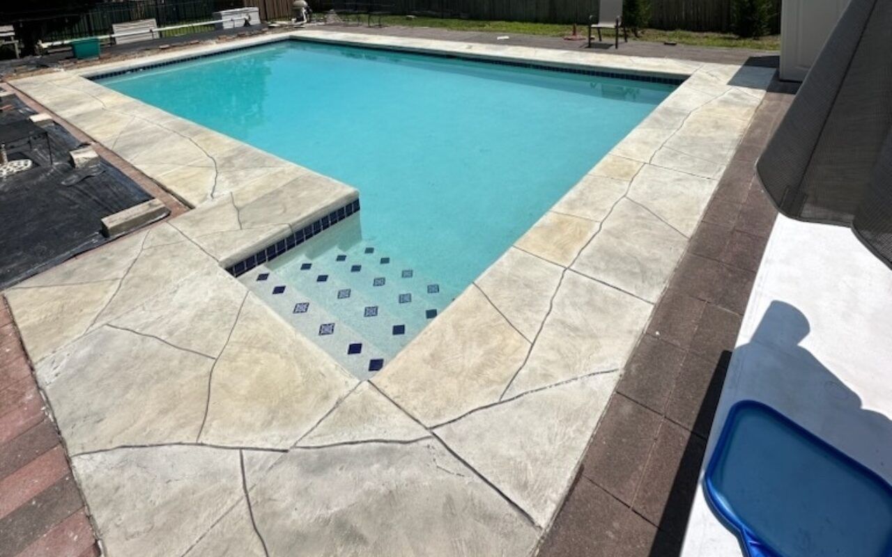 Outdoor swimming pool with light blue water and beige stone deck, viewed from the corner.