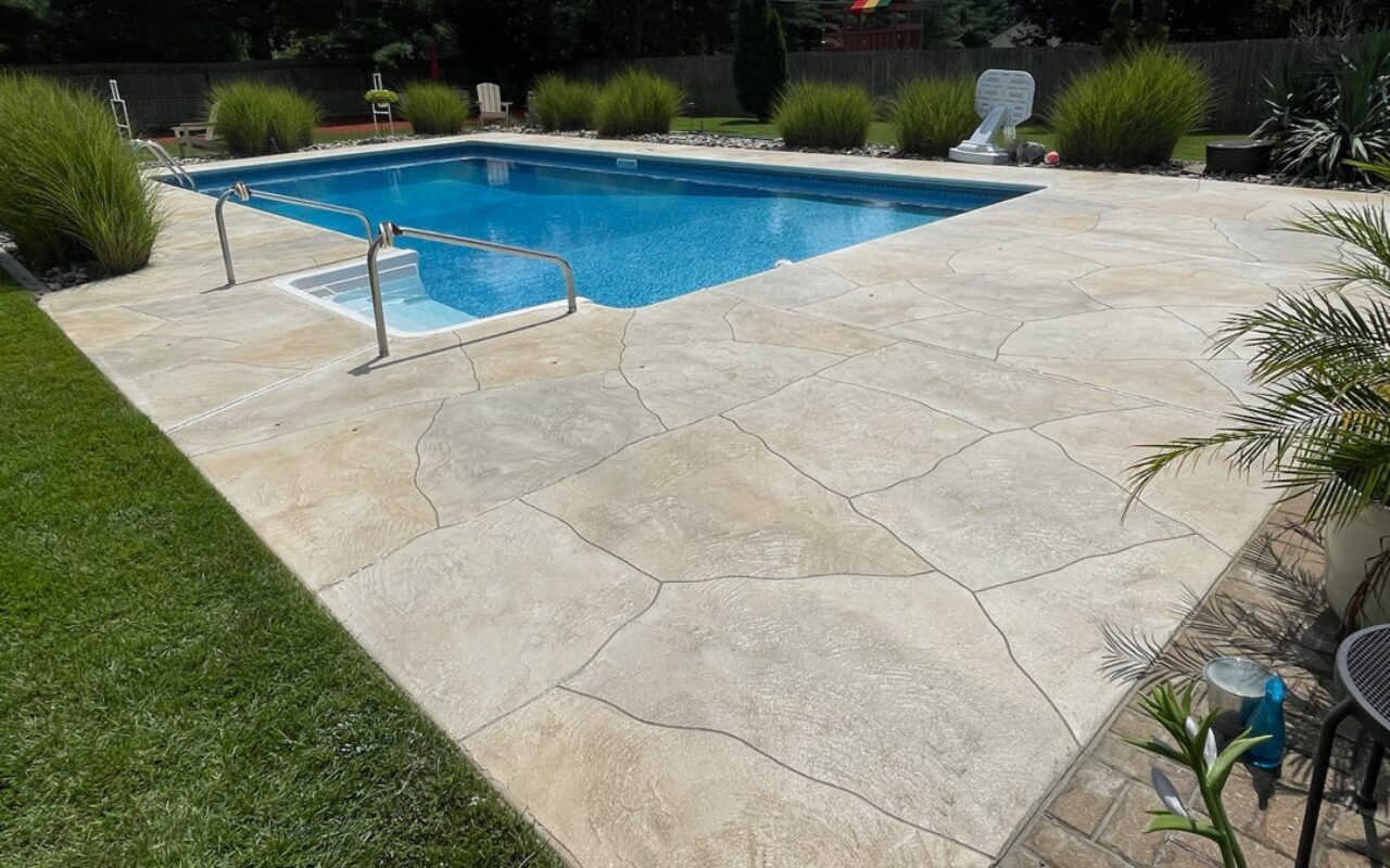 Backyard swimming pool with a tan stone patio, green lawn, and surrounding shrubs and lounge chair