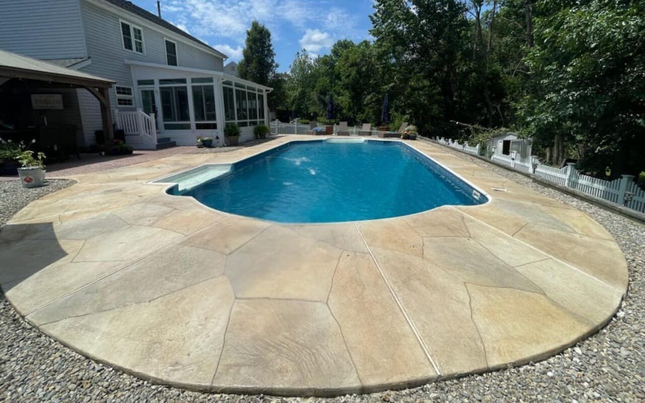 Backyard swimming pool beside a house, surrounded by a tan stone patio and trees.