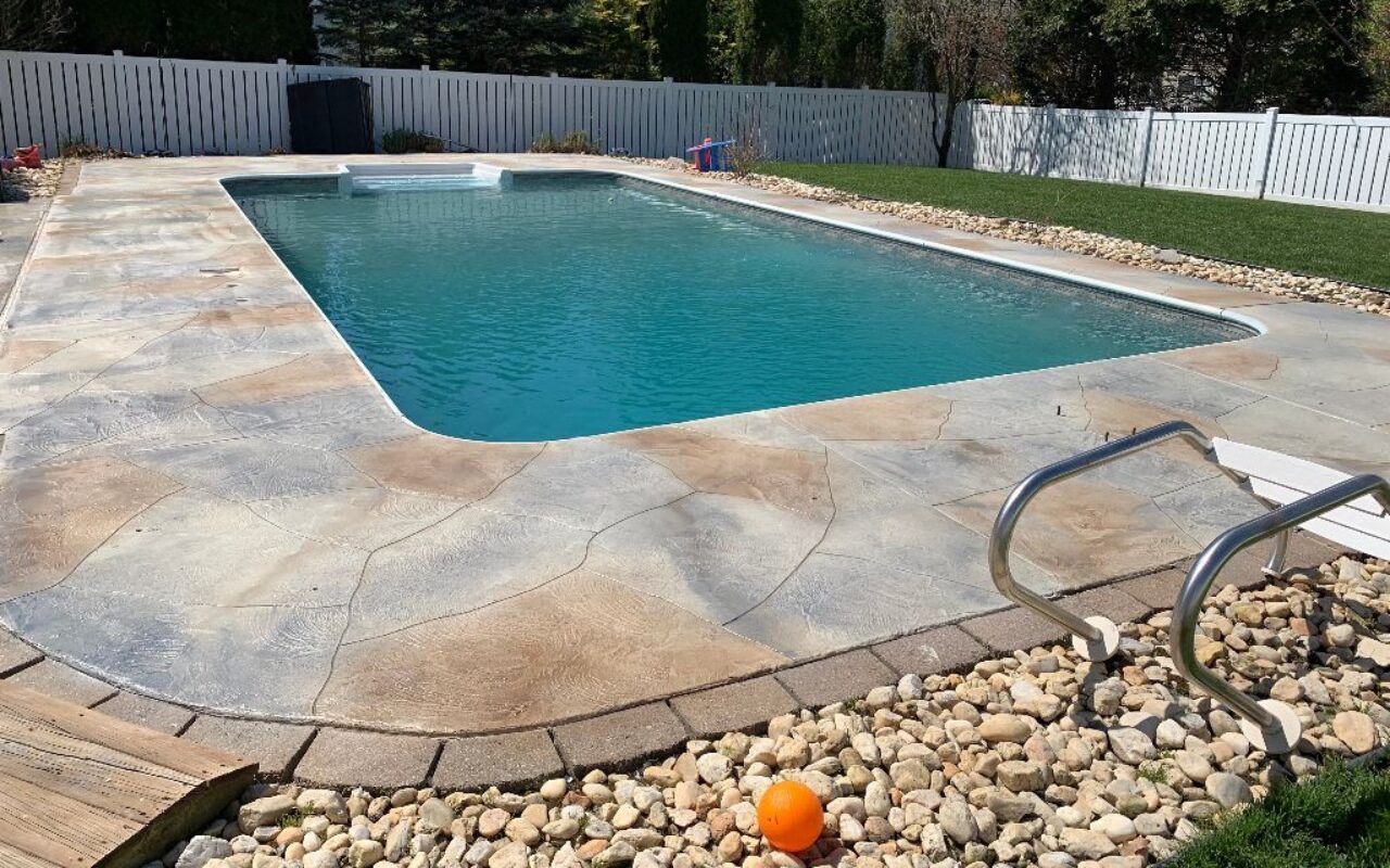 Backyard swimming pool with blue water, concrete deck, fence, and a lounge chair beside rocks and an orange ball