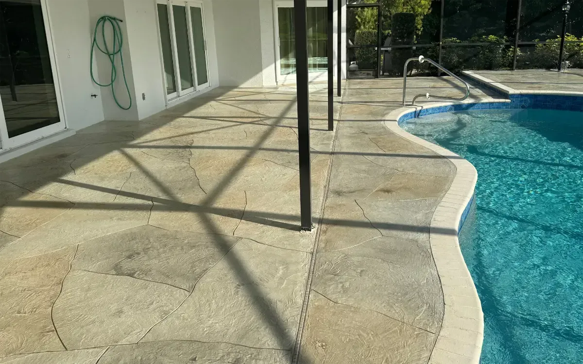 Patio beside a swimming pool with stone paving, house windows, and shadows from railing posts.