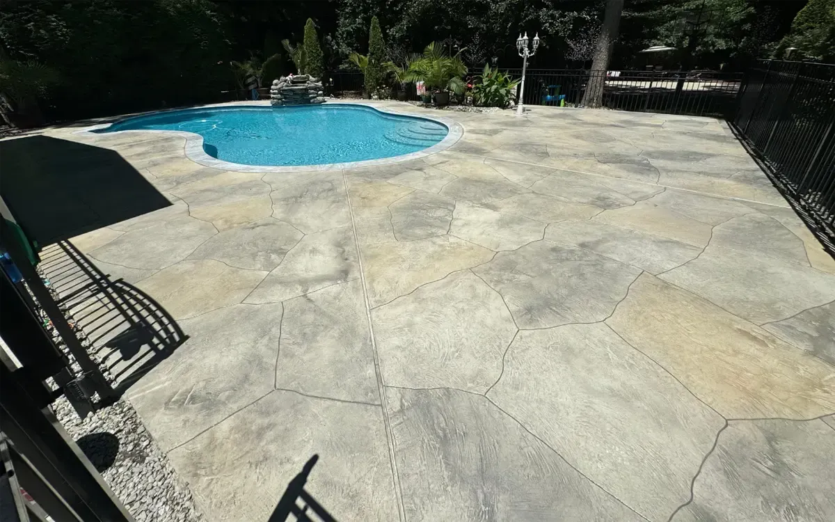 Backyard patio with a small blue swimming pool and a large stone deck in sunlight