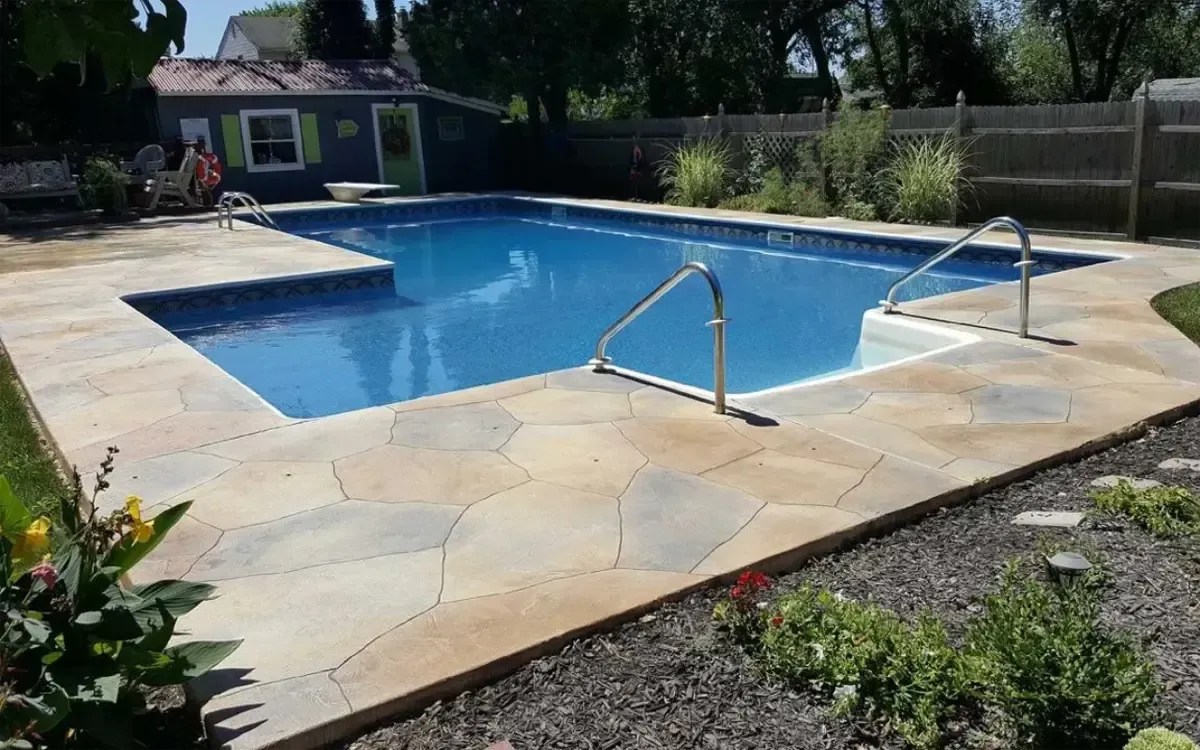Backyard swimming pool with beige stone deck and metal handrails, surrounded by greenery and fence