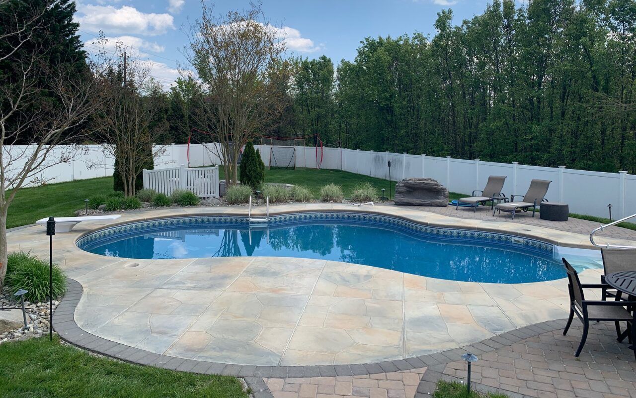 Backyard swimming pool with stone deck, lounge chairs, fire pit, and white fence surrounded by trees
