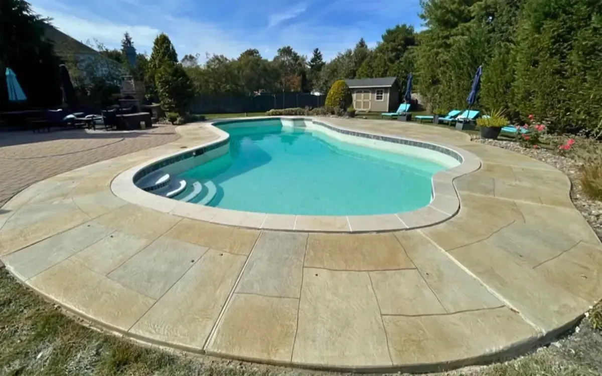 Backyard swimming pool with stone patio, turquoise water, and surrounding trees under a blue sky