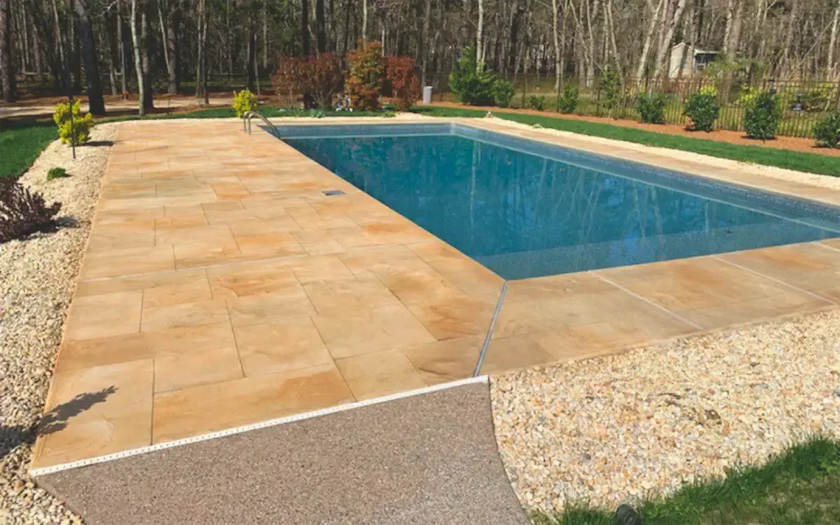 Rectangular blue in-ground pool with tan paver deck in a wooded backyard