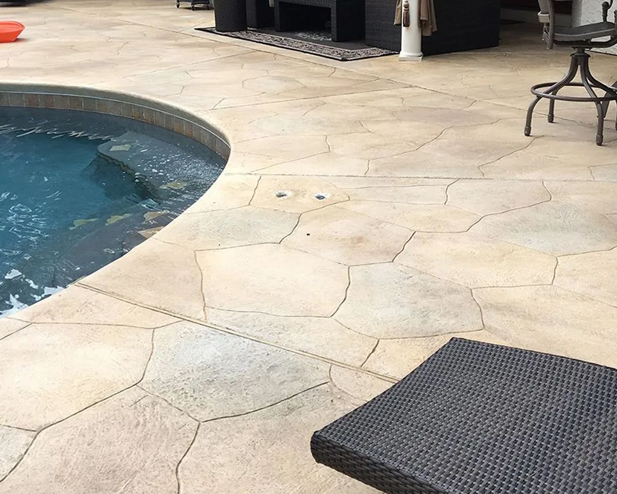 Poolside patio with curved blue pool, beige stone paving, and a dark outdoor mat.