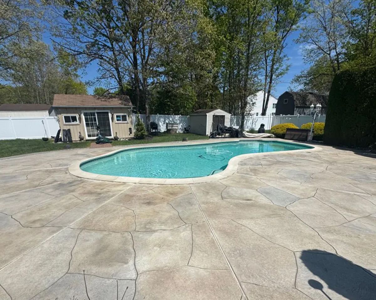 Backyard swimming pool with a light stone patio, trees, and buildings in the background under a clear blue sky