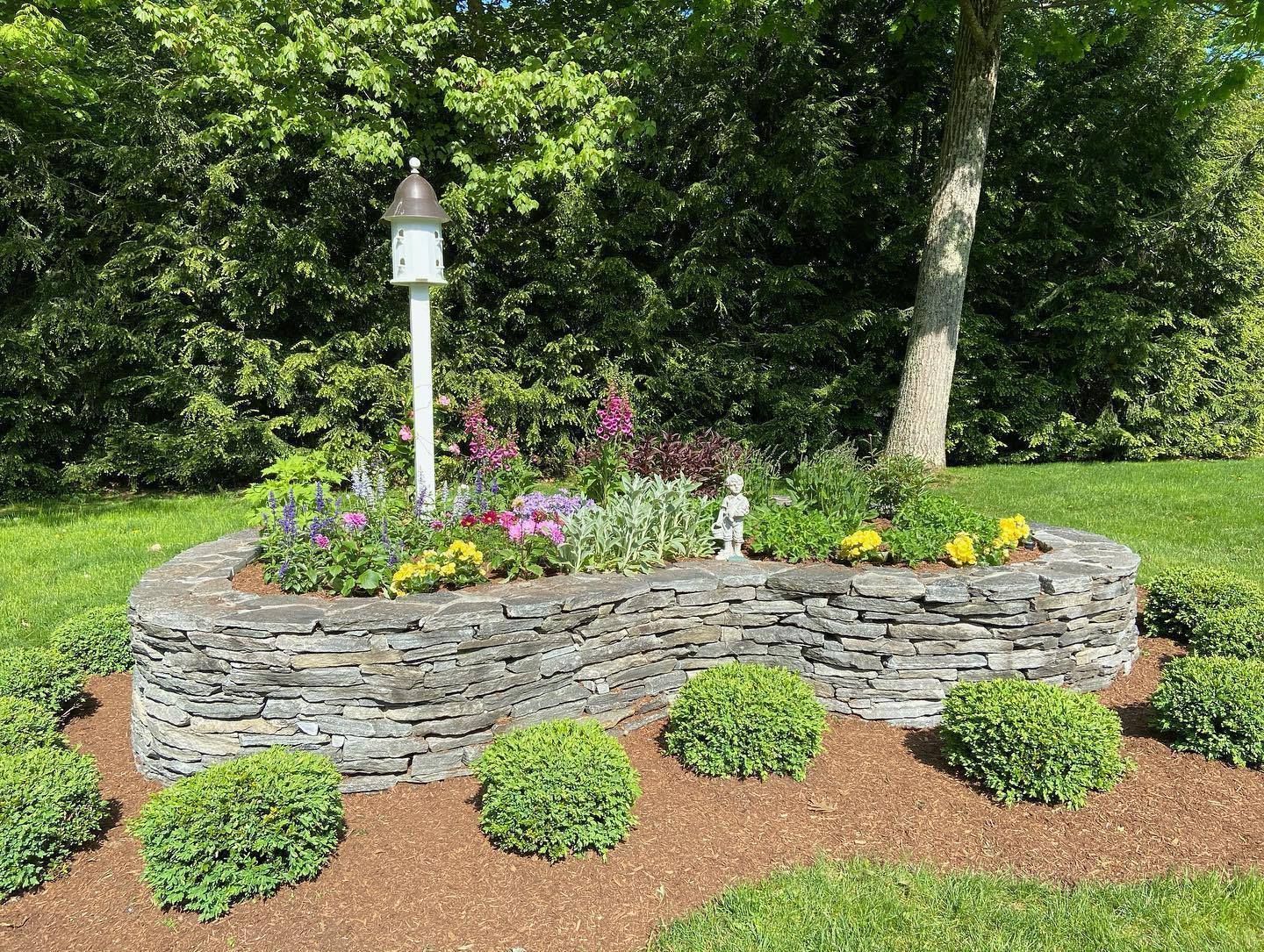 Stone-walled flower bed with blooming flowers, birdhouse, and shrubs on a grassy lawn.