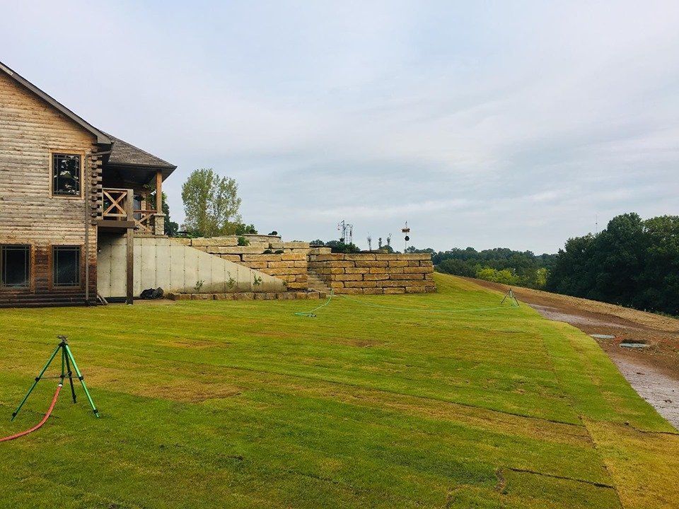 Wooden cabin with a grassy lawn, terraced retaining wall, and cloudy sky.
