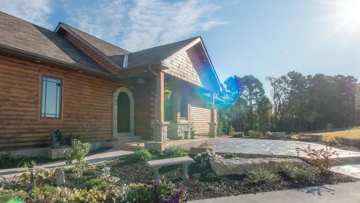 Wooden house exterior with a porch, sunny sky. Landscaping in front includes a stone patio.