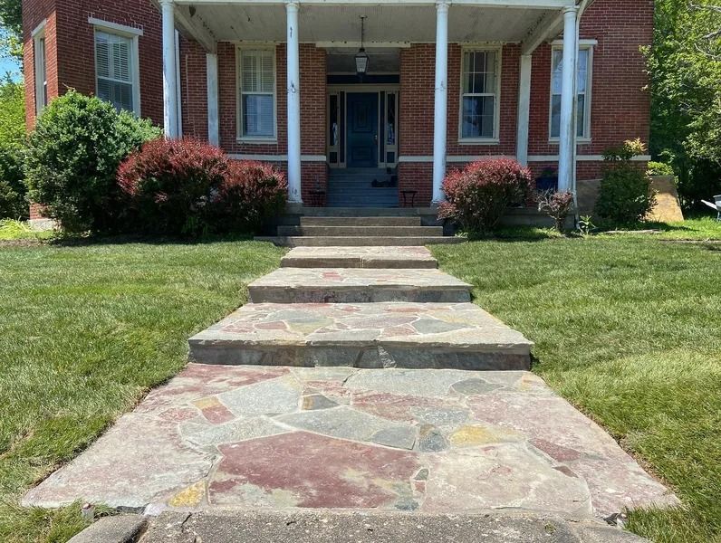 Stone walkway leading to a brick house with a porch. Green lawn and shrubbery.
