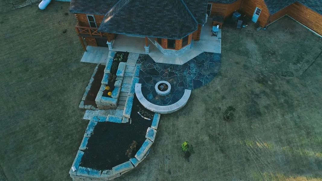 Aerial view of a house with a stone patio, fire pit, and landscaping on a grassy lot.