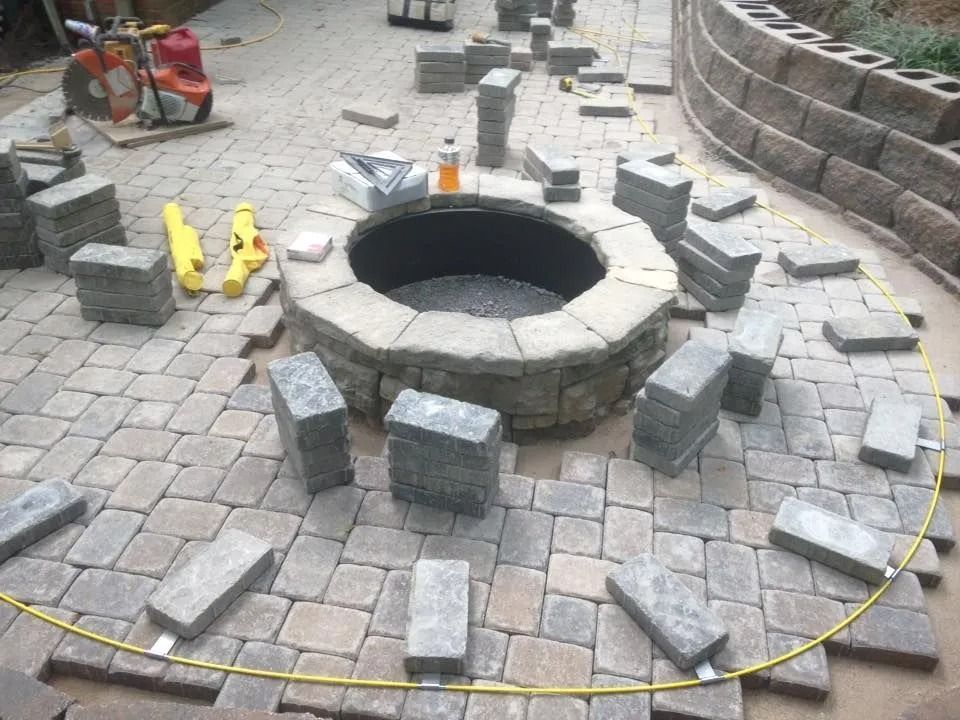 Paver patio under construction, with a fire pit, bricks, and a retaining wall.