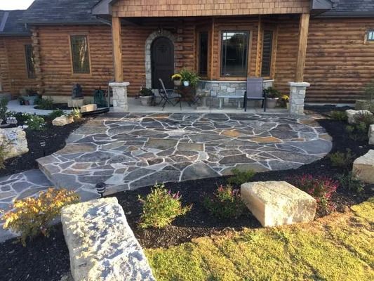 Stone patio in front of a log cabin with landscaping, including boulders, plants, and black mulch.