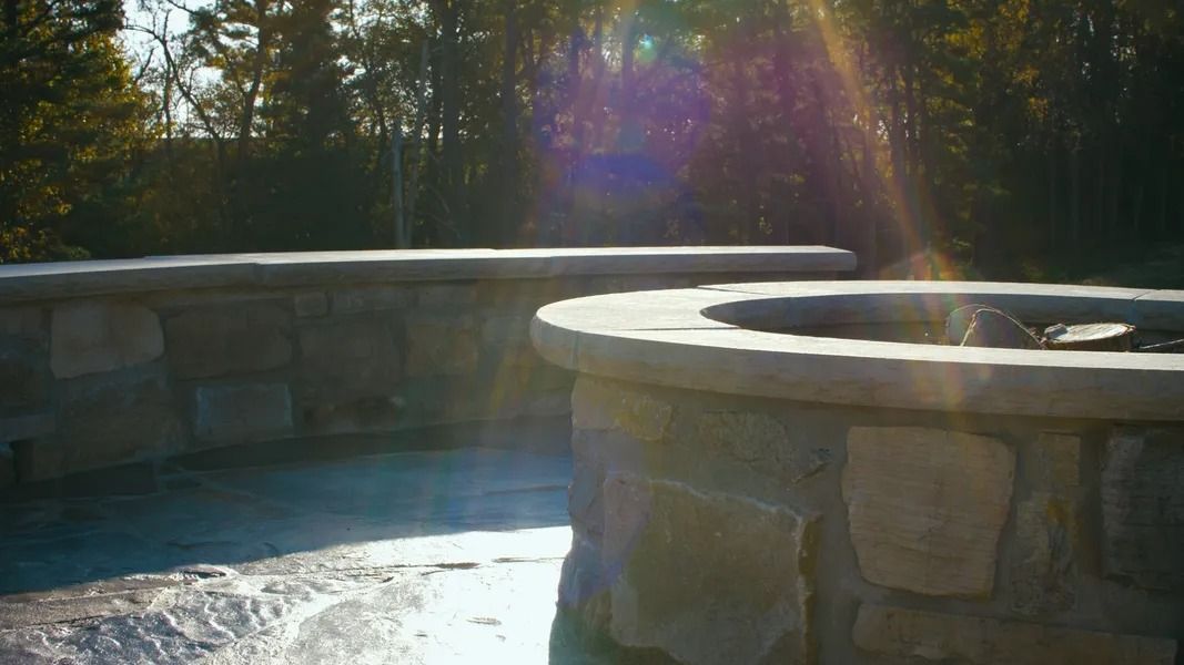 Stone fire pit and seating with sunlight, trees in the background.