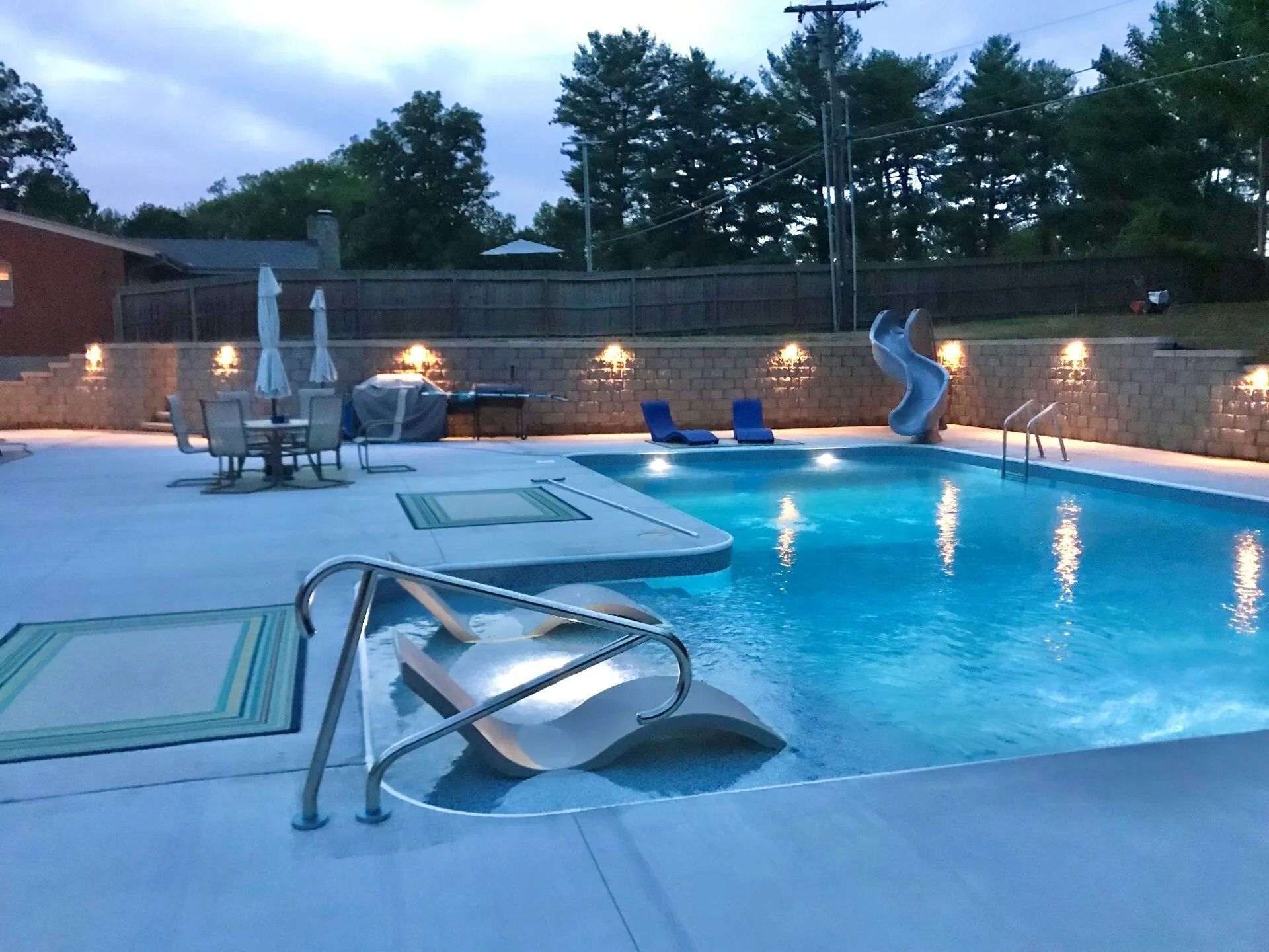 Pool area with illuminated pool, lounge chairs, dining table, and retaining wall.