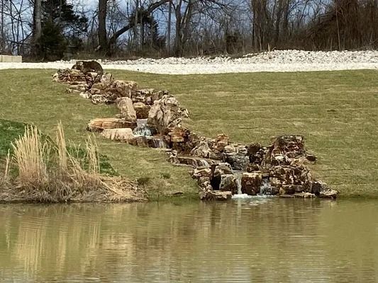 Waterfall flowing down a rocky hillside into a pond, surrounded by grass and trees.