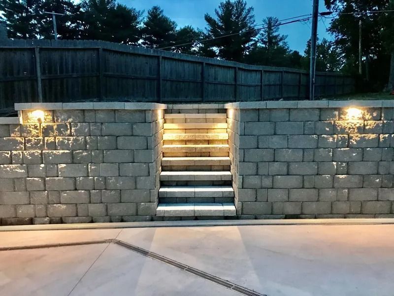 Stone retaining wall with lit steps leading up, flanked by lights, evening sky.