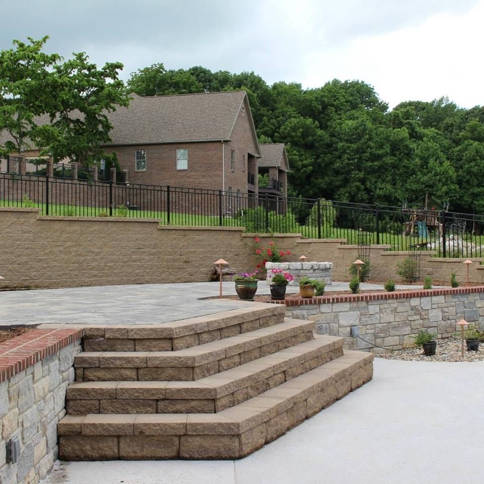 Stone steps lead up to a tiered paver patio and retaining wall outside a brick house, backed by a lush green forest.
