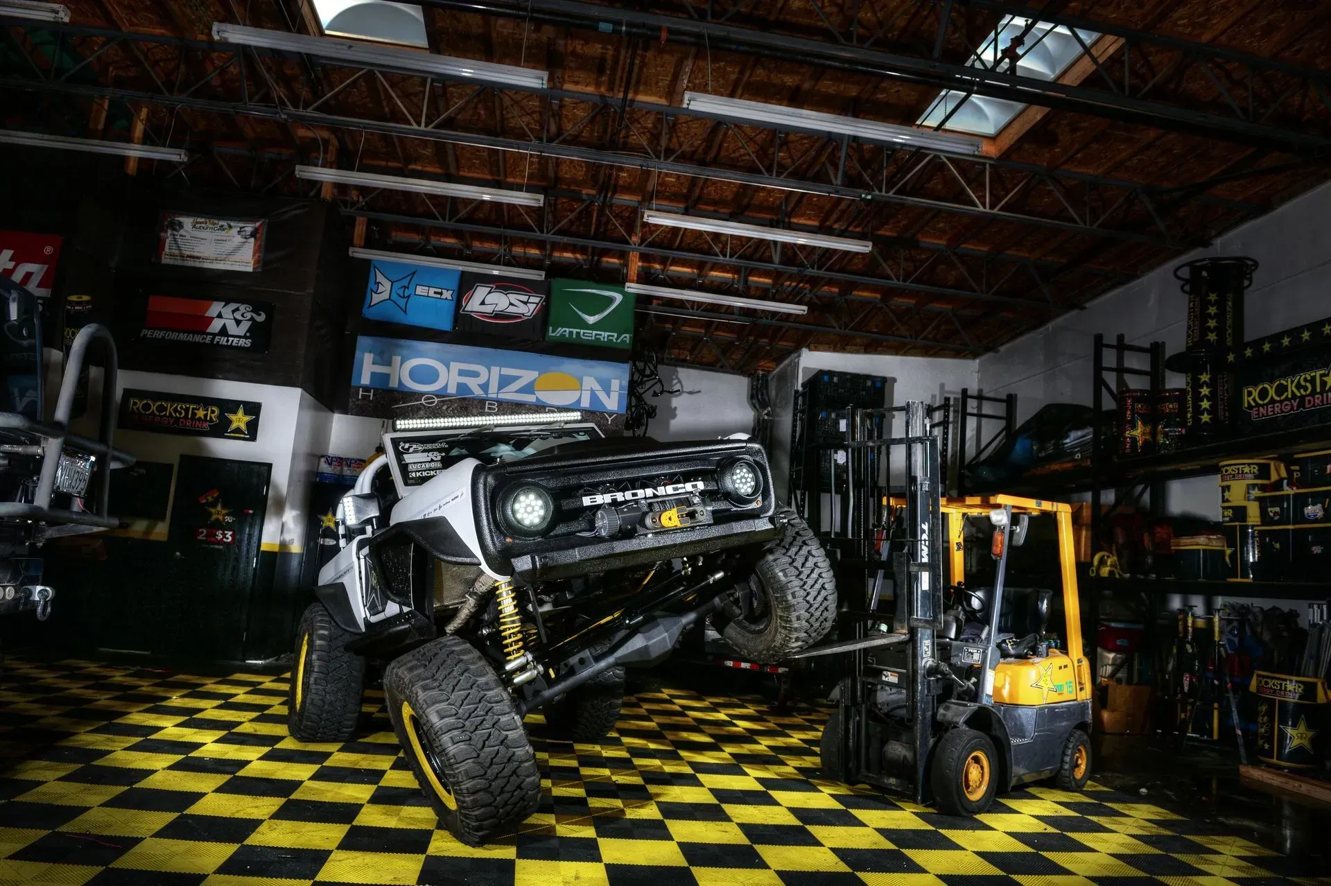 Off-road truck lifted by a forklift inside a workshop with checkered flooring, lit by overhead lights.