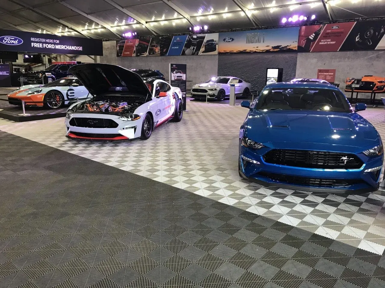 Ford Mustangs on display at an event. White, blue, and orange cars inside a tent.