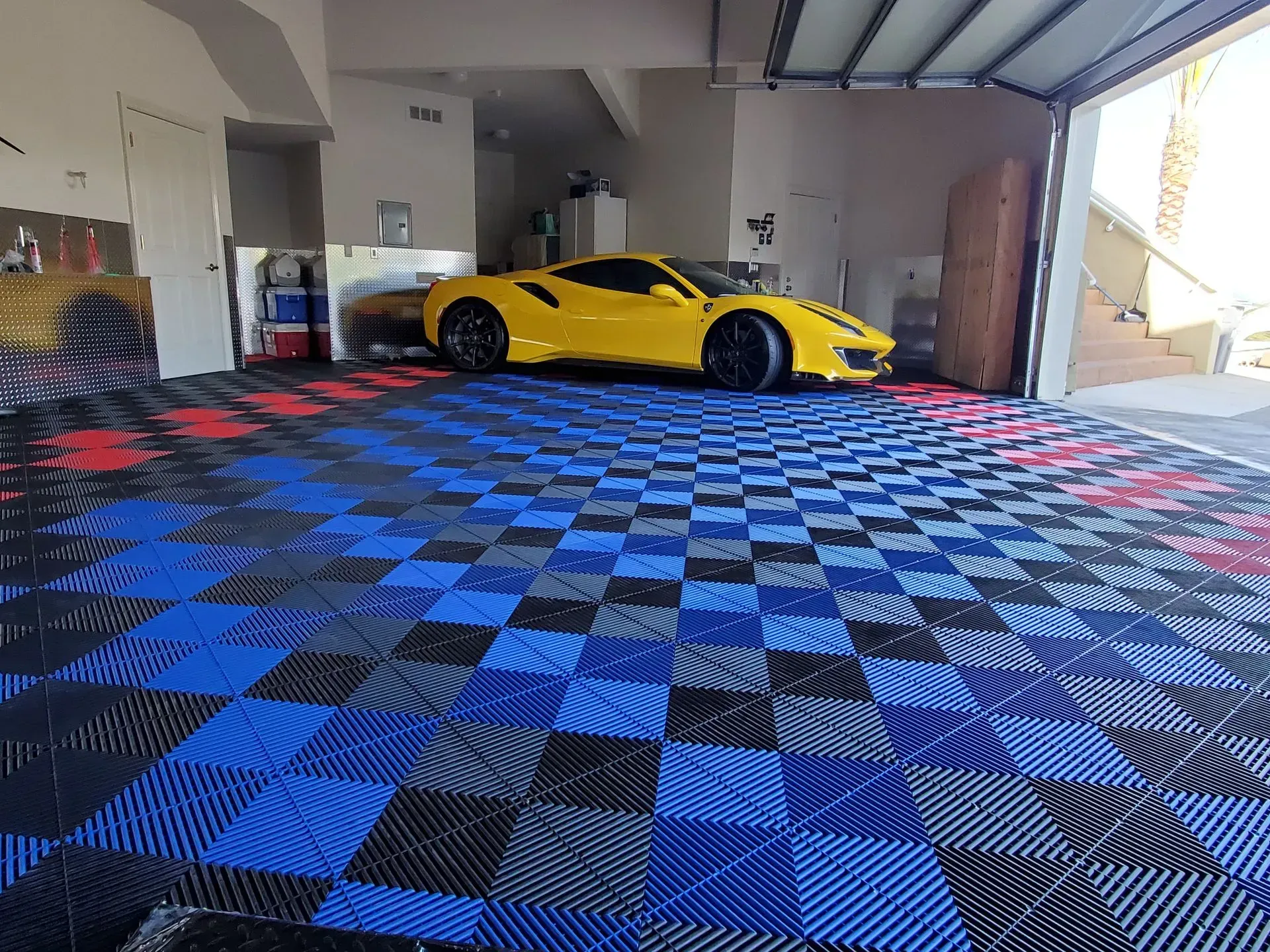 Yellow sports car in garage with blue, black, and red checkered floor tiles. Garage door open.