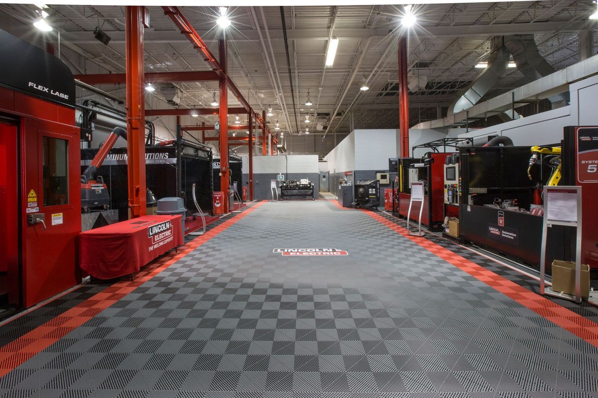 A trade show floor with machinery, booths, and a checkered floor. Red and black accents are visible.