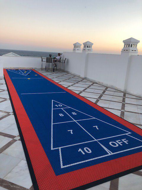 Shuffleboard court on a rooftop patio with two people seated nearby at dusk. Blue court with red border.