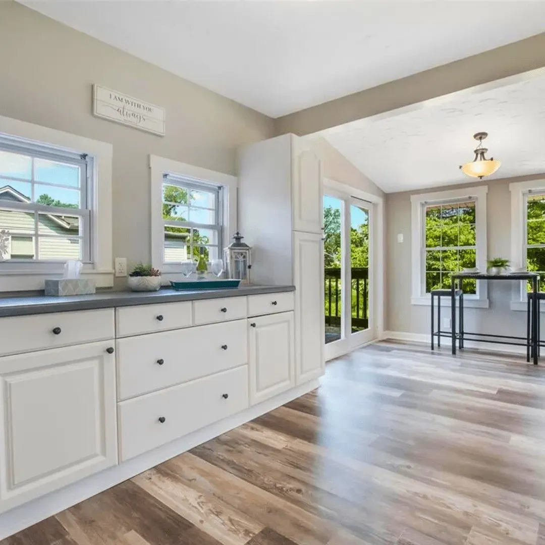 White kitchen cabinets and drawers, gray countertop, two windows, wood floor, open to dining area.