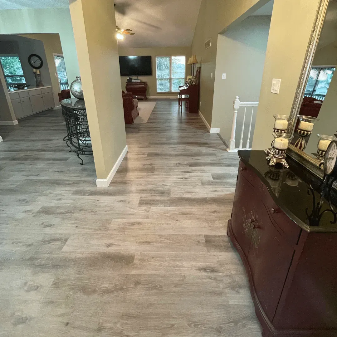 A hallway with wood-look flooring leads to a living room. A decorative cabinet and mirror are in the foreground.