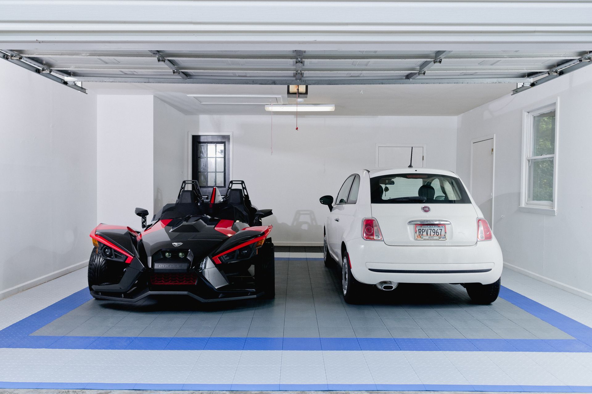 Two vehicles in a garage: a black three-wheeled Slingshot and a white Fiat 500. Blue floor outline.
