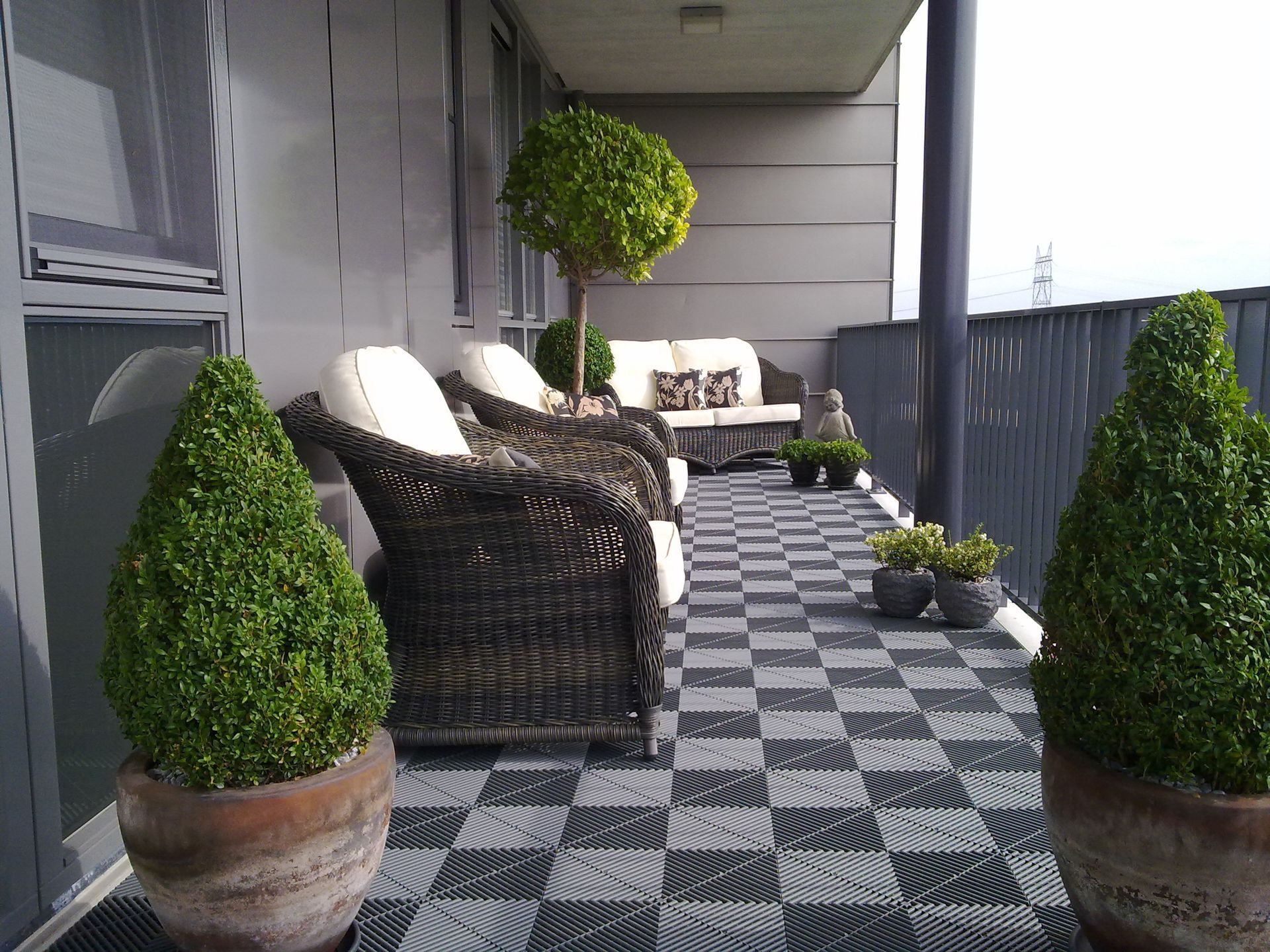 Balcony with gray checkerboard flooring, wicker furniture, potted topiaries, and a sofa.